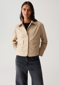 Giacca beige cropped con colletto, chiusura frontale a bottoni e due grandi tasche. Indossata sopra un top nero con texture e jeans scuri.
