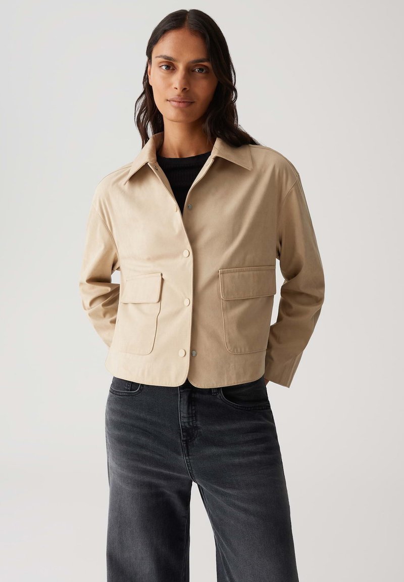 Giacca beige cropped con colletto, chiusura frontale a bottoni e due grandi tasche. Indossata sopra un top nero con texture e jeans scuri.