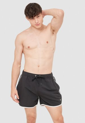 Joven de pie sin camiseta, con pantalones cortos de baño negros con cordón y ribete blanco, un brazo doblado detrás de la cabeza, sobre un fondo liso.
