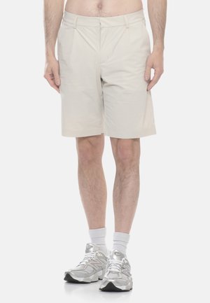 Uomo che indossa pantaloncini beige fino al ginocchio, calzini bianchi e scarpe da corsa argento, in piedi con le mani rilassate lungo i fianchi.