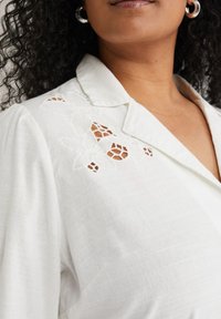 Camicia bianca con colletto, caratterizzata da ricami floreali e dettagli a taglio. Realizzata in un tessuto testurizzato con un leggero motivo a righe.