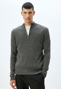 Suéter gris de punto acanalado con cuello de media cremallera y mangas largas. La tela parece suave con un acabado texturizado. Corte casual, usado sobre una camisa blanca.