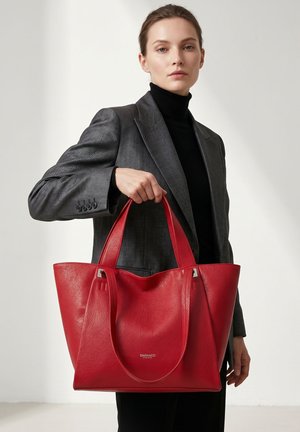 Femme en blazer gris et col roulé noir tenant un grand sac cabas en cuir rouge avec des doubles poignées.