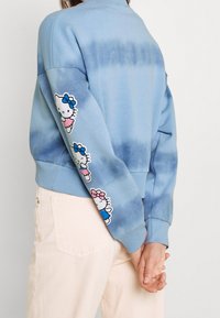 Světle modrý tie-dye svetr s žebrovanými manžetami a lemem. Na levém rukávu se nachází tři aplikace Hello Kitty v různých barvách.