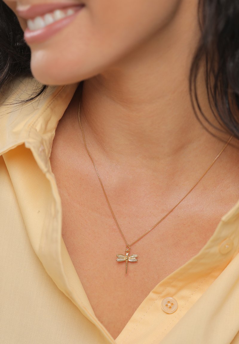 Zlatý náhrdelník s príveskom v podobe vážky s detailnými krídlami, nosený na svetložltej košeli s viditeľným gombíkom.