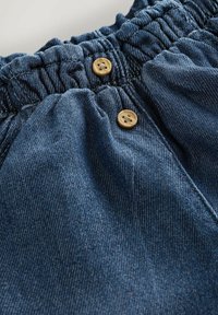 Farmerrövidnadrág elasztikus derékpánttal, két fa gombbal és finom kék varrással díszítve. Puha tapintású és kényelmes szabású.