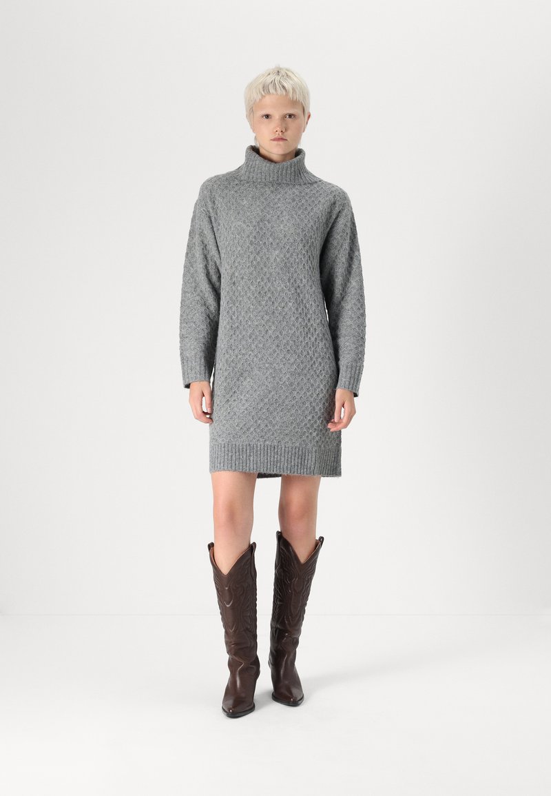 Robe en gris tricotée avec col roulé, dotée d'un motif en diamant texturé et d'un ourlet côtelé. Associée à des bottes de cow-boy marron jusqu'au genou.
