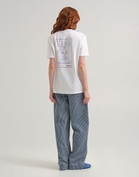 Persona con capelli rossi che indossa una maglietta bianca "Lee" con un personaggio dei cartoni animati e pantaloni larghi a righe blu, in piedi davanti a uno sfondo semplice.