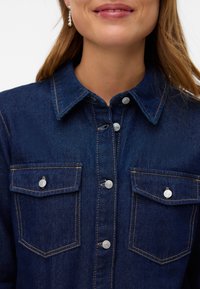 Femme souriante portant une chemise en jean bleu foncé avec des poches poitrine boutonnées et des boutons argentés, montrée des épaules au menton.