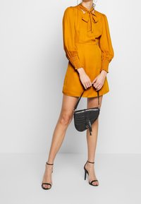 Senapsgul klänning med långa ärmar och en fluga vid halslinningen, kompletterad med en svart strukturerad axelväska och högklackade sandaler.