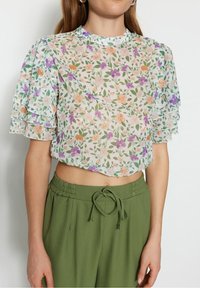 Blusa cropped con motivo floreale e maniche sovrapposte trasparenti in tonalità di rosa, viola e verde, abbinata a pantaloni olive green a vita alta.