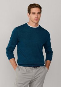 Ciemnoniebieski sweter z długimi rękawami i okrągłym dekoltem, w zestawie z jasnoszarymi spodniami. Gładka faktura i ściągacze na rękawach.