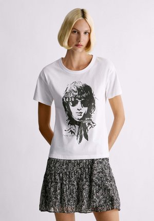 T-shirt bianca in cotone con stampa grafica nera di una persona con occhiali da sole e testo, abbinata a una gonna nera con motivo.