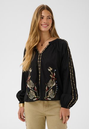 Femme aux longs cheveux blonds portant un chemisier noir avec broderie florale et un pantalon beige, souriante et regardant sur le côté sur un fond uni.