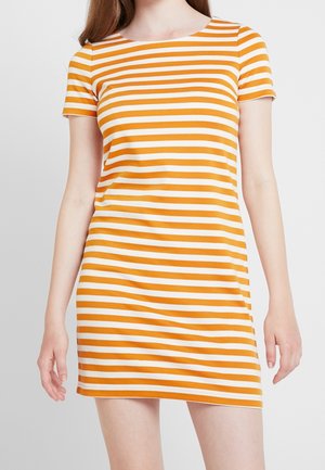 Kobieta w sukience z krótkim rękawem sięgającej do kolan, z poziomymi pomarańczowo-białymi pasami, stojąca na tle jasnego, jednolitego tła.