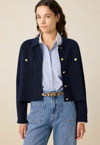 Marineblauwe gebreide cardigan met gouden knopen en twee borstzakken, gedragen over een lichtblauw gestreept shirt, gecombineerd met denim jeans.