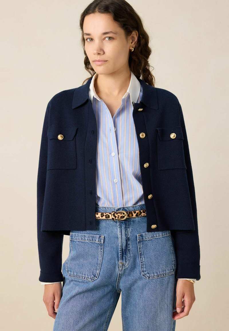 Marineblauwe gebreide cardigan met gouden knopen en twee borstzakken, gedragen over een lichtblauw gestreept shirt, gecombineerd met denim jeans.