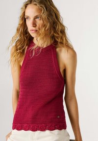 Vrouw met lang, krullend blond haar, gekleed in een mouwloos, gestructureerd donkerroze gebreid topje en lichtgekleurde broek, kijkt iets naar links.