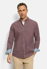 Mann trägt ein rot-blau kariertes Hemd mit hellblauen umgeschlagenen Ärmeln und beigen Hosen, steht vor einem weißen Hintergrund.