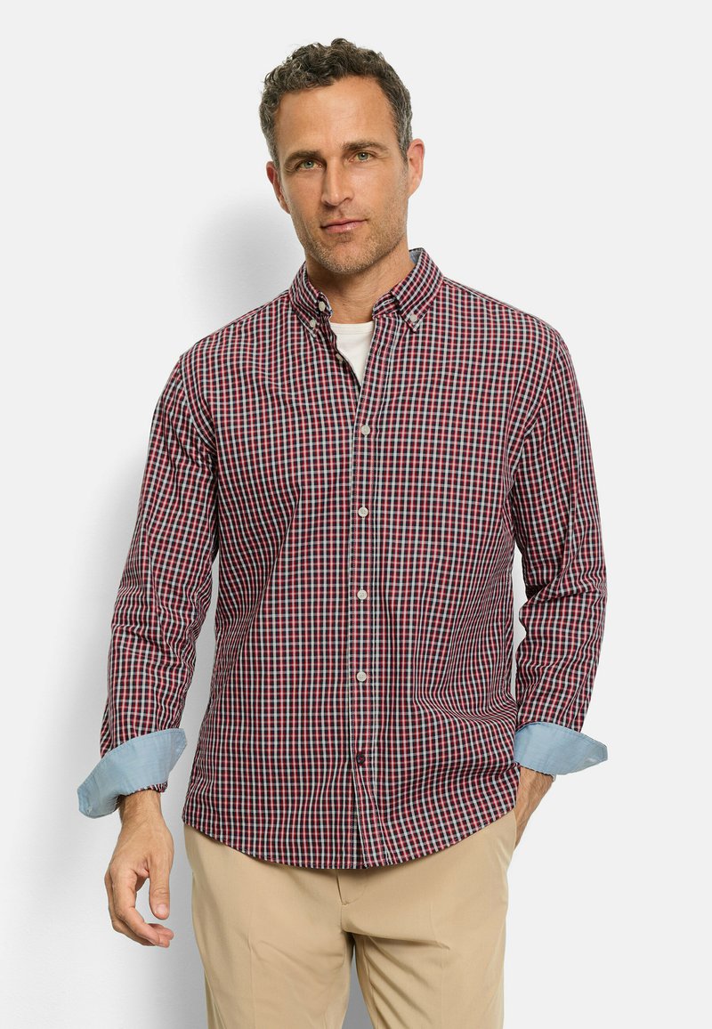 Mann trägt ein rot-blau kariertes Hemd mit hellblauen umgeschlagenen Ärmeln und beigen Hosen, steht vor einem weißen Hintergrund.