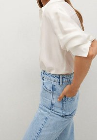 Femme portant une chemise blanche à manches longues retroussées et un jean taille haute bleu clair, avec la main dans la poche arrière.