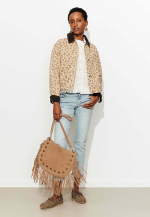 Femme portant une veste beige à fleurs, un chemisier blanc, un jean bleu clair et des mocassins marron, tenant un sac à main beige à franges sur un fond blanc.