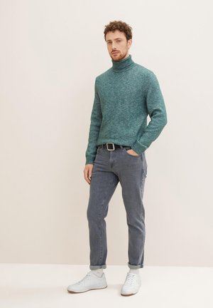 Teal wool turtleneck sweater, ribbed collar; paired with slim grey jeans and white sneakers. Standing posture against a neutral background.