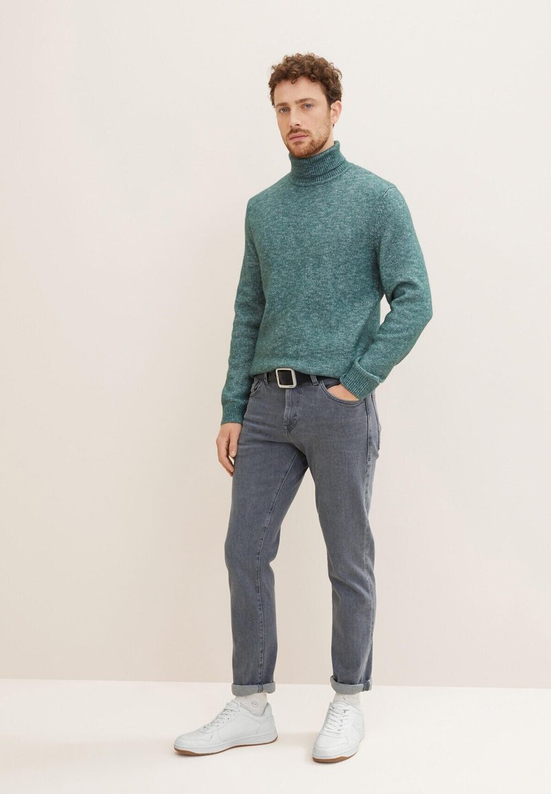 Teal wool turtleneck sweater, ribbed collar; paired with slim grey jeans and white sneakers. Standing posture against a neutral background.