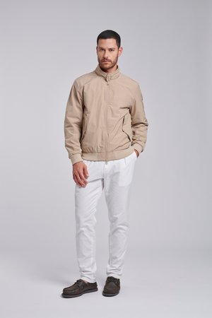 Man met baard die een beige jas, witte broek en donkerbruine schoenen draagt, staand tegen een effen lichtgrijze achtergrond.