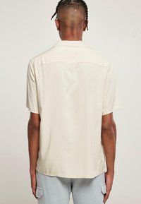 Personne avec des cheveux tressés portant une chemise beige à manches courtes et un pantalon cargo gris clair, vue de dos contre un fond uni.