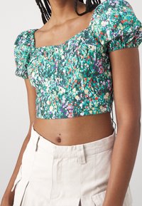 Zelený květinový crop top s nabíranými rukávy, s čtvercovým výstřihem a zkrčenou texturou. V kombinaci s světlými šortkami s vysokým pasem.