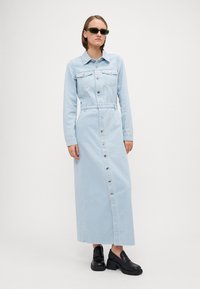 Svetlomodrá džínová košeľa s dlhými rukávmi, s gombíkovým zapínaním vpredu a dvoma náprsními vreckami. Kombinovaná s čiernymi robustnými topánkami a slnečnými okuliarmi.