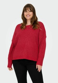 Maglione rosso lavorato a uncinetto con una vestibilità ampia, maniche larghe e un motivo testurizzato. L'orlo inferiore è leggermente sfrangiato per un aspetto rilassato.