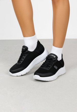 Piernas con calcetines blancos hasta el tobillo y zapatillas deportivas negras con suelas blancas, de pie sobre un suelo gris claro con un fondo liso.