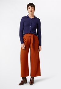 Marineblauer gerippter Cardigan mit Knopfverschluss, kombiniert mit weiten rostfarbenen Hosen mit gebundenem Bund, ergänzt durch glänzende braune Ankle Boots.