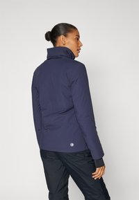 Navy blue jacket with a high collar, showing a smooth outer texture. Features a logo on the back and dark pants complementing the look.