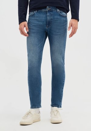 Uomo che indossa jeans blu slim fit, calzini bianchi e sneakers bianche, in piedi con le mani rilassate lungo i fianchi su uno sfondo chiaro uniforme.