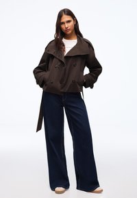 Giovane donna in piedi con le mani nelle tasche, indossa un trench corto marrone scuro, una camicia bianca, jeans ampi scuri e scarpe beige.