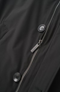 Schwarze Jacke mit einer glatten, matten Oberfläche. Enthält einen stabilen Reißverschluss mit einem gebrandeten Zipper und einem großen schwarzen Knopf.