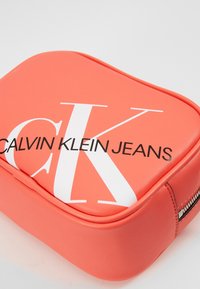 Korallfärgad läderpung med rundade kanter, prydd med en framträdande vit logotyp "CALVIN KLEIN JEANS" och silvrig dragkedjedetalj. Slät textur.