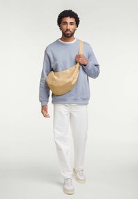 Borsa a tracolla beige realizzata in tessuto liscio, con forma arrotondata, tracolla regolabile e dettagli in oro, indossata sopra un maglione blu.
