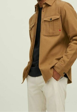 Mężczyzna ubrany w jasno-brązową kurtkę koszulową zapinaną na guziki, na czarnej koszulce, z lewą ręką w kieszeni białych spodni.