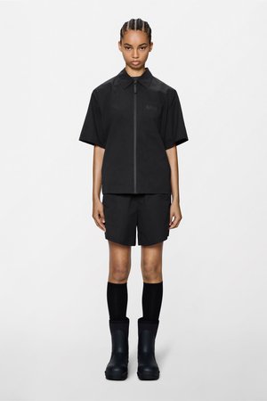 Frau steht nach vorne gerichtet, trägt ein schwarzes kurzärmeliges Reißverschluss-Shirt, passende Shorts, schwarze knielange Socken und schwarze Stiefel vor weißem Hintergrund.