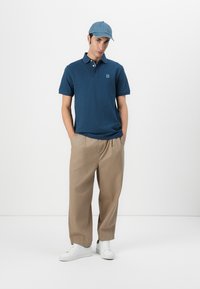 Ναυτικό πόλο μπλούζα με μικρό λογότυπο, συνδυασμένη με ανοιχτό μπεζ παντελόνι και λευκά αθλητικά παπούτσια. Το μοντέλο φοράει ένα ανοιχτό μπλε καπέλο.