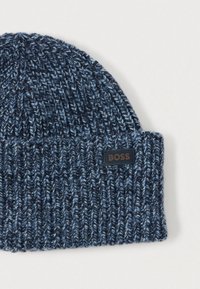 Πλεκτό σκουφάκι σε σκούρο μπλε με ανοιχτόχρωμες μπλε κηλίδες· υφή με ανάγλυφο μοτίβο· διαθέτει μαύρη ετικέτα με το λογότυπο 'BOSS' στο διπλωμένο μέρος.