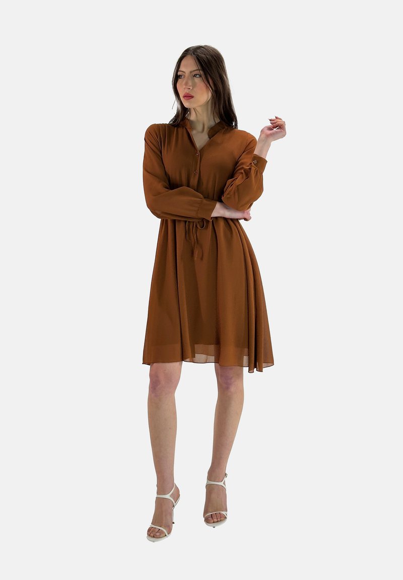 Vestido marrón, hasta la rodilla, con botones y mangas largas. Hecho de tela ligera, tiene un cinturón atado y un corte holgado. Combinado con sandalias.