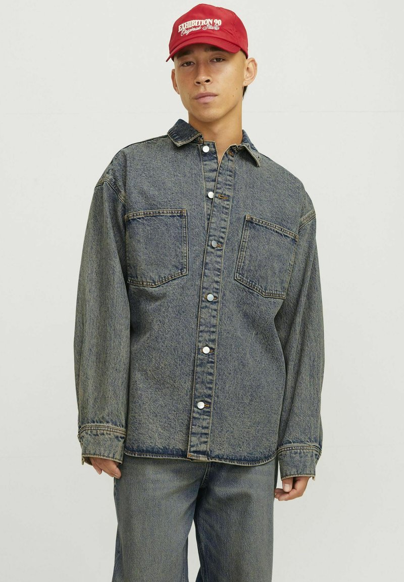 Camicia di denim blu sbiadito con due tasche sul petto, chiusura a bottoni e maniche lunghe, abbinata a pantaloni di denim coordinati e un cappellino rosso.