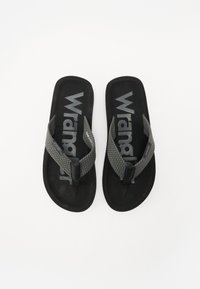 Svarte flip-flops med vevde grå stropper og strukturert svart såle. Har et fremtredende "Wrangler"-logo på innersålen.