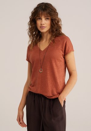 Frau mit mittellangem, lockigem Haar, die ein rostfarbenes T-Shirt mit V-Ausschnitt, schwarze Hosen, eine Anhänger-Halskette trägt und die Hand in der Tasche hat, vor einem einfarbigen Hintergrund.