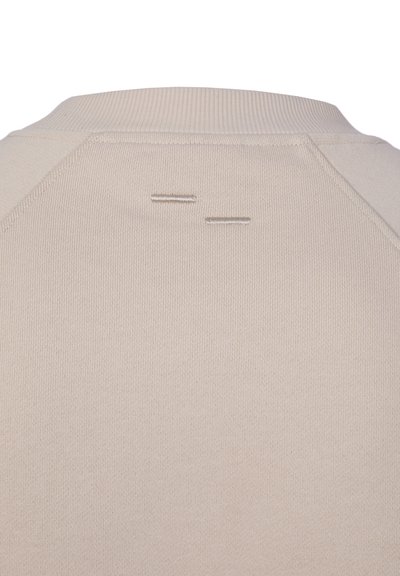 Detail béžové látky zobrazující zadní část výstřihu oděvu s žebrovaným límcem a dvěma malými vodorovnými prošitými detaily.